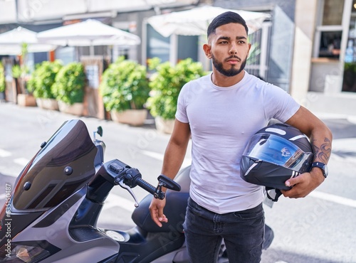 Young latin man holding helmet standing by motorbike at street