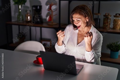 Middle age hispanic woman using laptop at home at night very happy and excited doing winner gesture with arms raised, smiling and screaming for success. celebration concept.