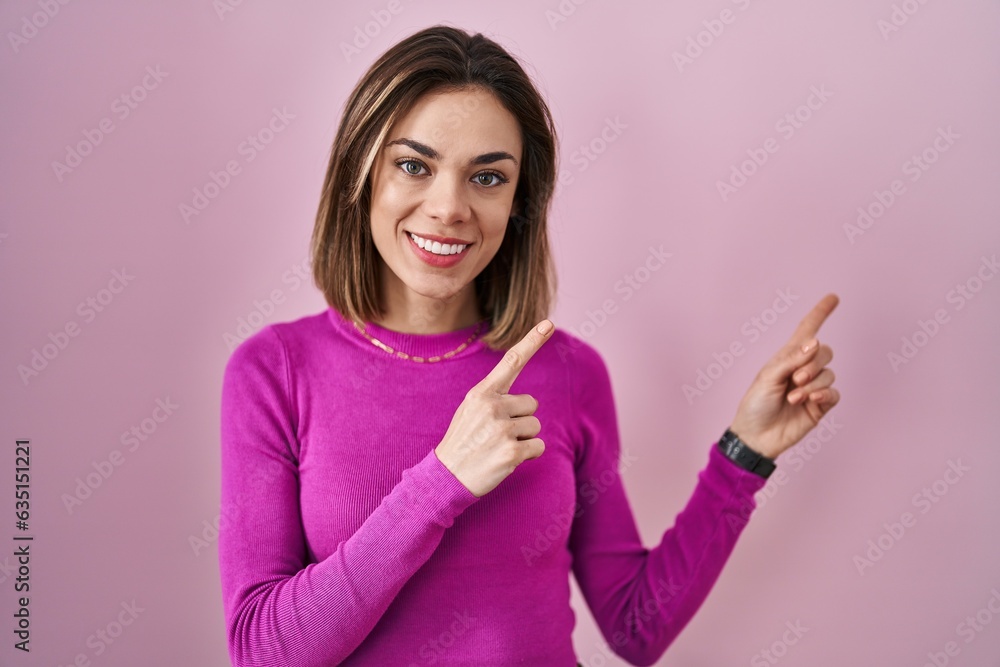 Hispanic woman standing over pink background smiling and looking at the camera pointing with two hands and fingers to the side.