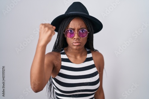 Young african american with braids wearing hat and sunglasses angry and mad raising fist frustrated and furious while shouting with anger. rage and aggressive concept.