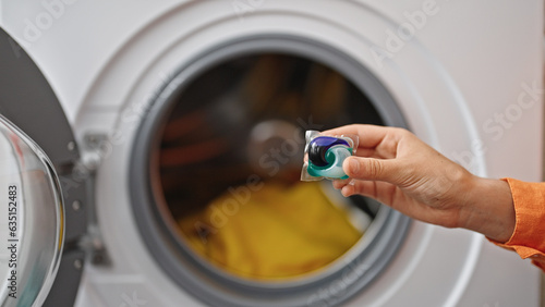 Young beautiful hispanic woman washing clothes holding detergent bag at laundry room