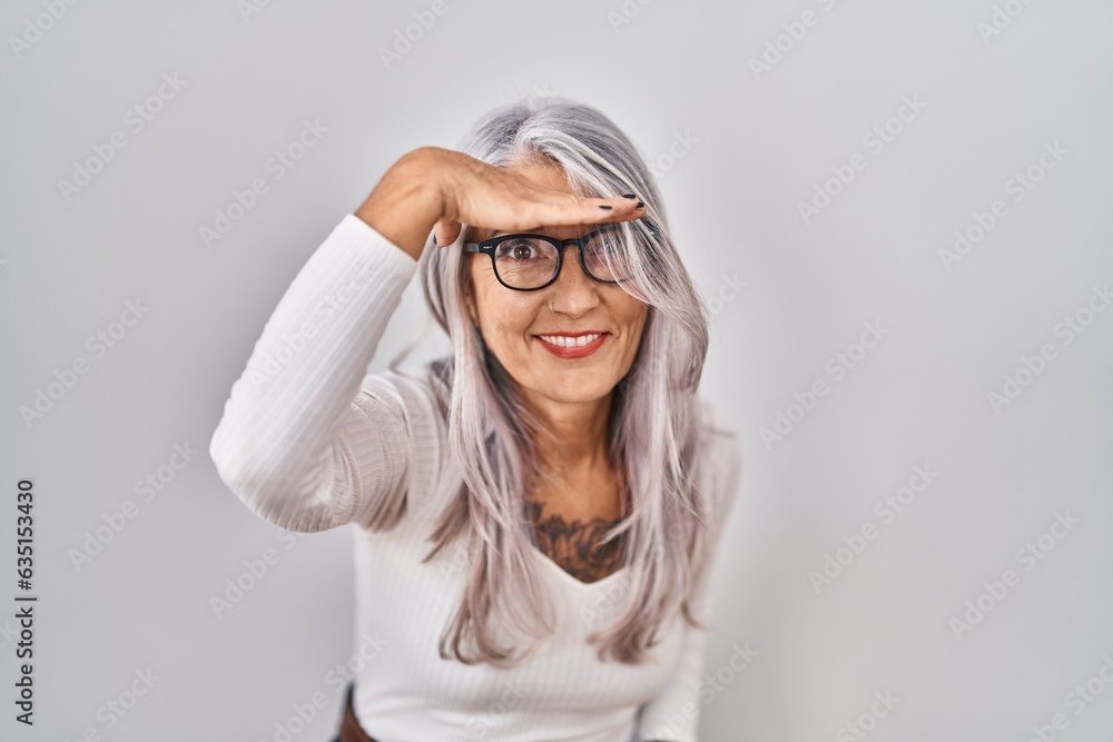 Middle age woman with grey hair standing over white background very happy and smiling looking far away with hand over head. searching concept. Middle age woman with grey hair standing over white background very happy and smiling looking far away with hand over head. searching concept.