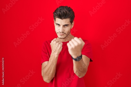 Young hispanic man standing over red background ready to fight with fist defense gesture, angry and upset face, afraid of problem