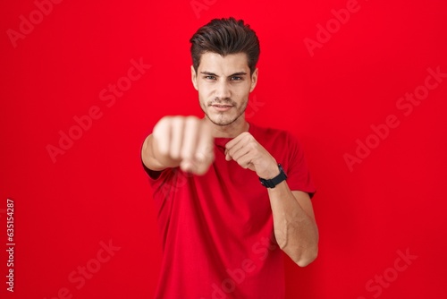 Young hispanic man standing over red background punching fist to fight, aggressive and angry attack, threat and violence