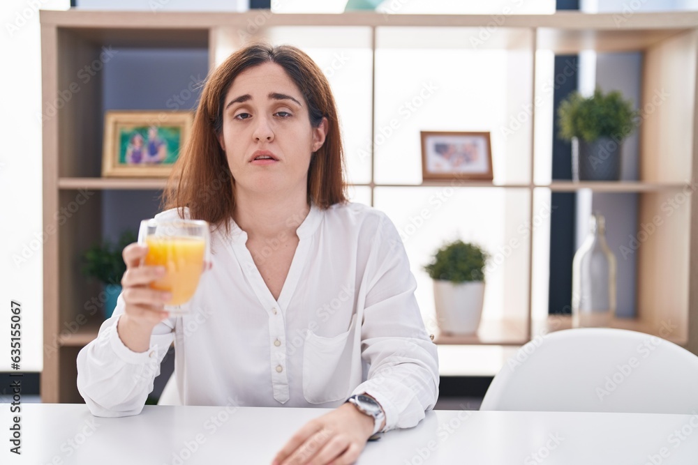 Brunette woman drinking glass of orange juice looking sleepy and tired, exhausted for fatigue and hangover, lazy eyes in the morning.
