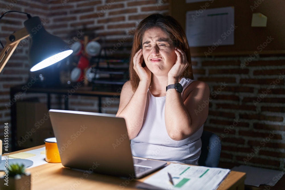 Brunette woman working at the office at night covering ears with fingers with annoyed expression for the noise of loud music. deaf concept. Brunette woman working at the office at night covering ears with fingers with annoyed expression for the noise of loud music. deaf concept.