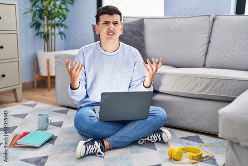 Non binary person studying using computer laptop sitting on the floor crazy and mad shouting and yelling with aggressive expression and arms raised. frustration concept. photo