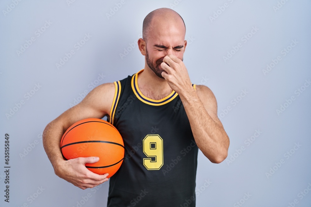 Young bald man with beard wearing basketball uniform holding ball smelling something stinky and disgusting, intolerable smell, holding breath with fingers on nose. bad smell