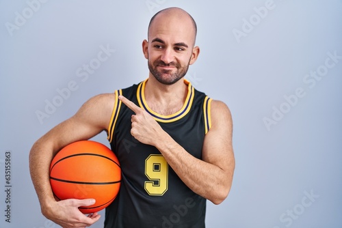 Young bald man with beard wearing basketball uniform holding ball pointing with hand finger to the side showing advertisement, serious and calm face