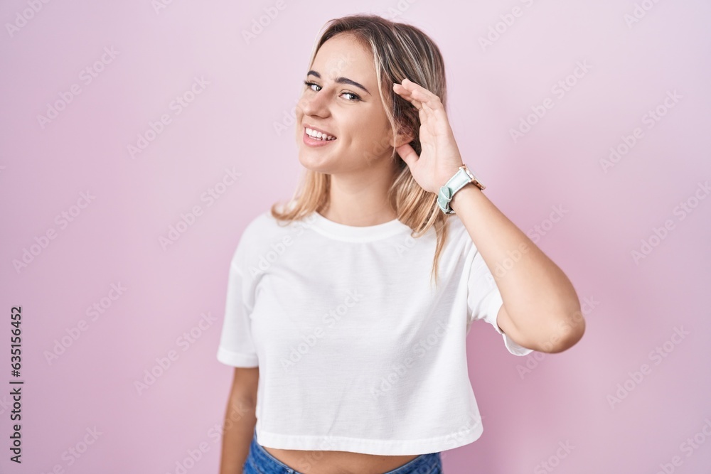 Young blonde woman standing over pink background smiling with hand over ear listening an hearing to rumor or gossip. deafness concept. Young blonde woman standing over pink background smiling with hand over ear listening an hearing to rumor or gossip. deafness concept.