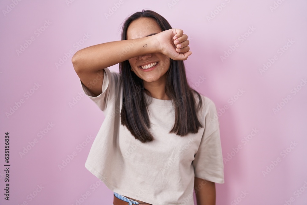 Young hispanic woman standing over pink background covering eyes with arm smiling cheerful and funny. blind concept. Young hispanic woman standing over pink background covering eyes with arm smiling cheerful and funny. blind concept.