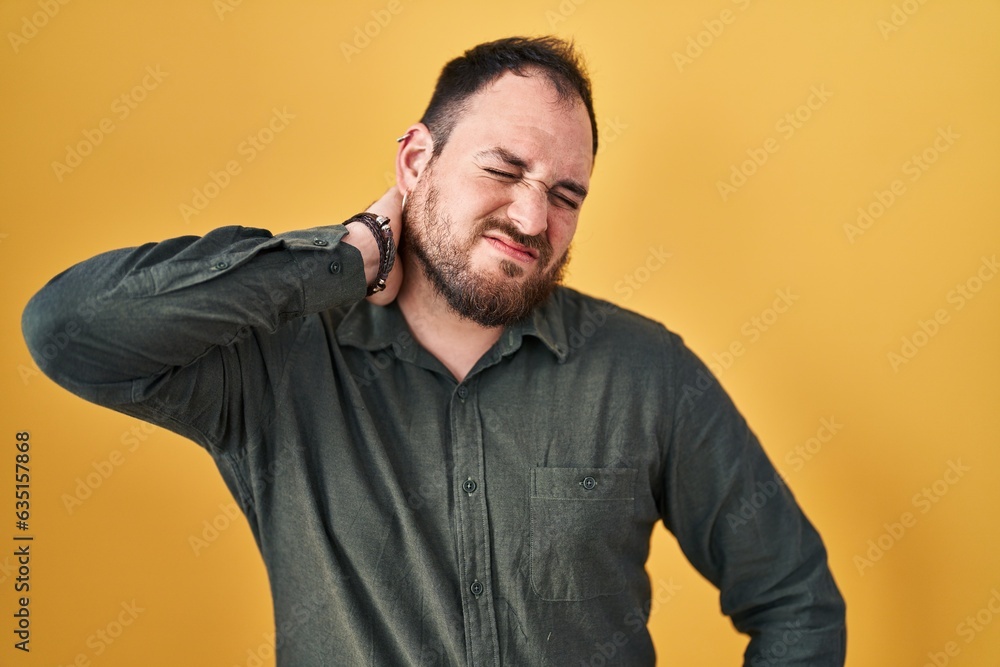 Plus size hispanic man with beard standing over yellow background suffering of neck ache injury, touching neck with hand, muscular pain Plus size hispanic man with beard standing over yellow background suffering of neck ache injury, touching neck with hand, muscular pain