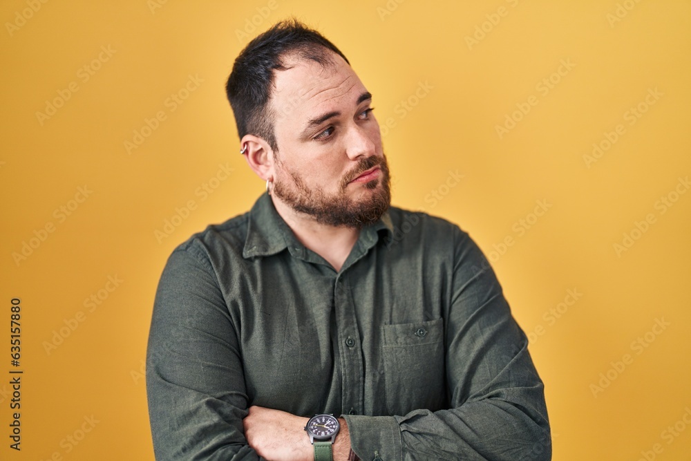 Fototapeta premium Plus size hispanic man with beard standing over yellow background looking to the side with arms crossed convinced and confident