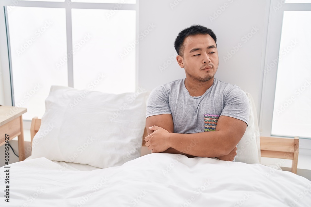 Young chinese man sitting on bed with unhappy expression and arms crossed gesture at bedroom Young chinese man sitting on bed with unhappy expression and arms crossed gesture at bedroom
