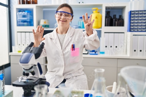 Hispanic girl with down syndrome working at scientist laboratory showing and pointing up with fingers number nine while smiling confident and happy.