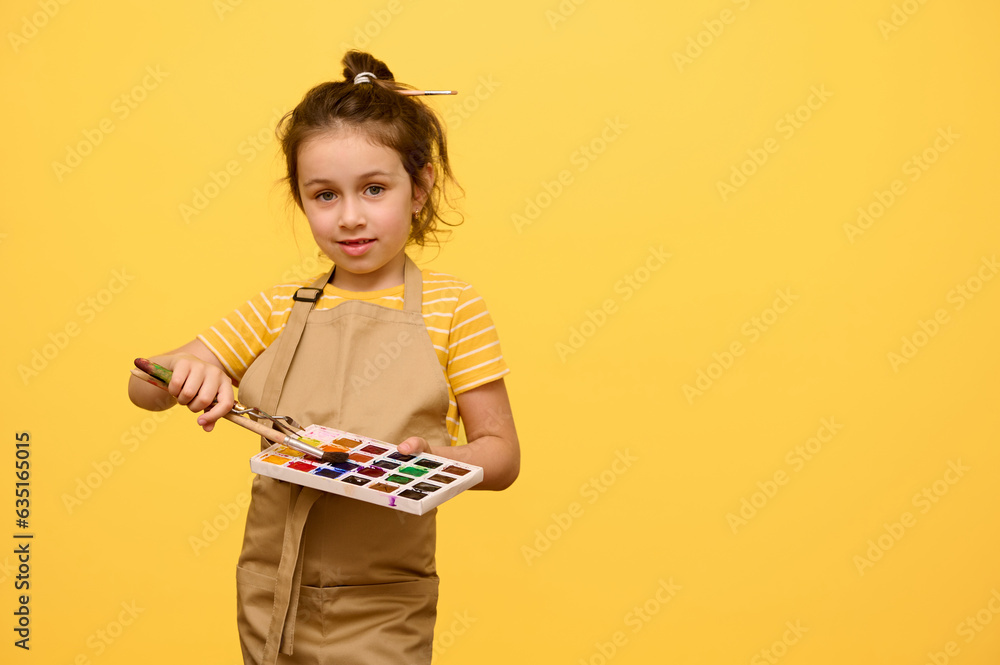 Adorable talented little kid girl painter artist in apron with paintbrush in hairstyle, holds watercolor palette and painting tools, isolated on yellow. Kids. Hobbies and leisures. Fine art concept Adorable talented little kid girl painter artist in apron with paintbrush in hairstyle, holds watercolor palette and painting tools, isolated on yellow. Kids. Hobbies and leisures. Fine art concept