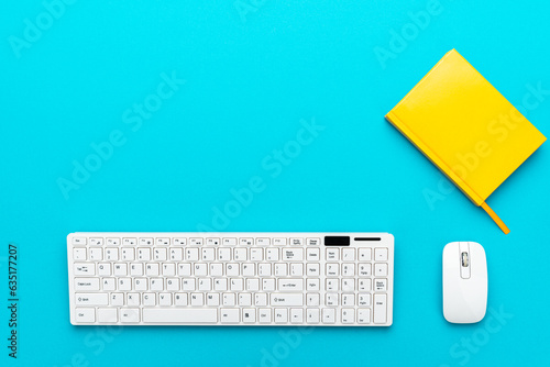 View from above on the office table with copy space. Minimalist photo of computer keyboard and mouse and notepad. Flat lay image of white computer peripherals over turquoise blue background.
