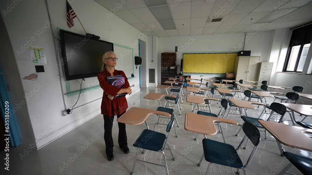 Wide angle view of sad unhappy teacher in large empty classroom with the lights turned off during budget cuts or a pandemic wondering about the future of education. Wide angle view of sad unhappy teacher in large empty classroom with the lights turned off during budget cuts or a pandemic wondering about the future of education.