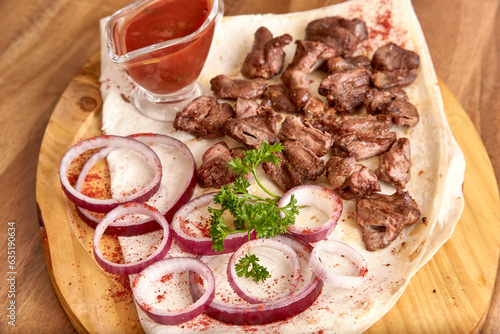 Part of the wooden board with small pieces of fried beef meat with red onion rings, sauce and pita bread on the wooden table, close-up perspective view, shallow depth of field. Meat and onion in focus