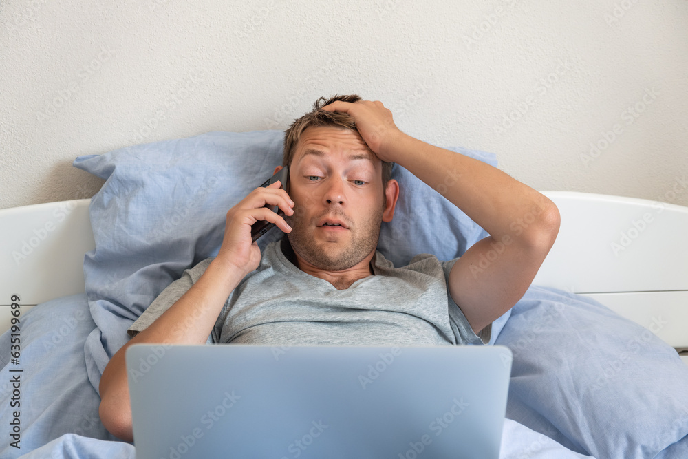 Upset disappointed man with mobile phone disputing looking at laptop while laying in a bed at home and solving online computer problem with technical support by smartphone Upset disappointed man with mobile phone disputing looking at laptop while laying in a bed at home and solving online computer problem with technical support by smartphone