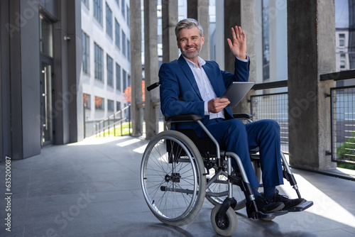 Happy business man in wheelchair with tablet using mobile device for work outdoors, waving hand.