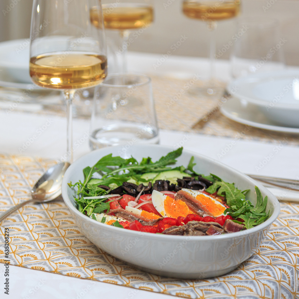 nicoise salad layered in a deep plate served with a knife and fork with a glass of water and wine