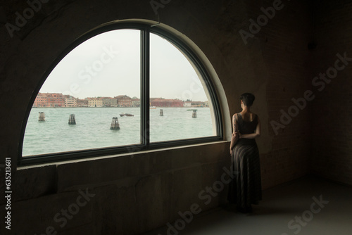 Una giovane donna sola osserva malinconica un canale di Venezia da una grande finestra panoramica ad arco photo