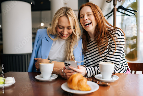 Portrait of two cheerful beautiful women friends sitting in cafe indoors, looking at phone and talking, laughing. Happy pretty females girlfriends using social media on smartphone in coffee shop.