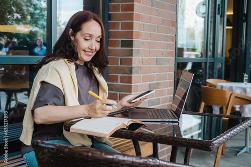 Middle aged woman working online in cafe outdoor writng in planner