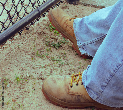 Legs of a person wearing dirty work boots and jeans on dirt next to a fence.
