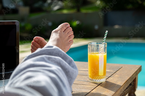 Close nos pés de uma mulher em cima da mesa à beira da piscina, com calças de moletom cinza e um copo de suco de laranja com canudo listrado em lindo dia ensolarado em fundo desfocado.
