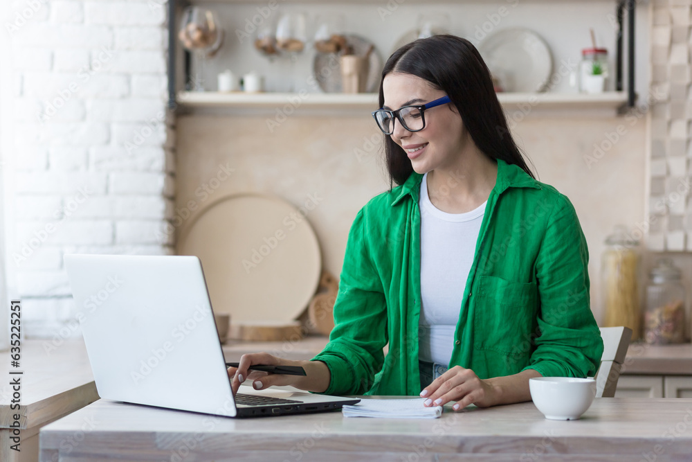 A young woman works and studies at home using a laptop. Sitting in the kitchen at the table and writing in a notebook. A young woman works and studies at home using a laptop. Sitting in the kitchen at the table and writing in a notebook.