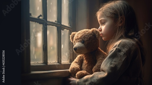 Sad looking orphan girl by the window holding a teddy bear photo