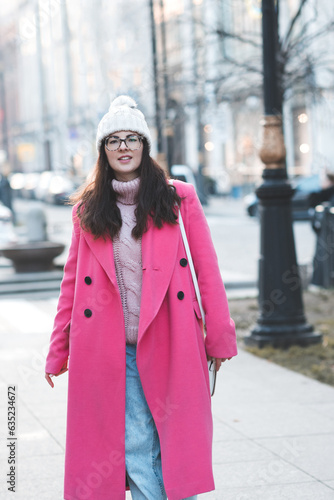 Happy smiling stylish woman wear pink winter jacket and knitted hat walk on city street outdoor. Fall season.