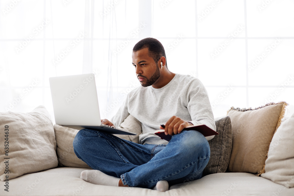 Focused young african american man in earphone using laptop during online education and holding notebook on couch at home Focused young african american man in earphone using laptop during online education and holding notebook on couch at home