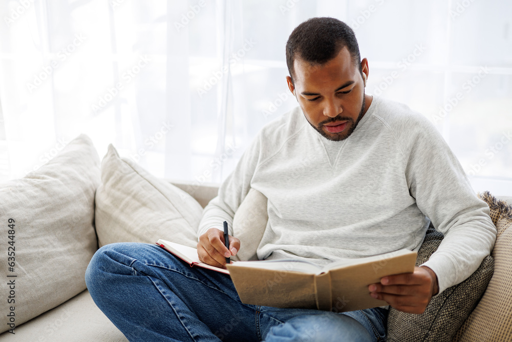 Young african american man in earphones reading book and writing on notebook on couch at home, self-education concept