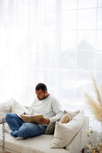 Young african american man in earphone writing on notebook and holding book on couch at home, self-education concept