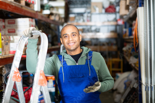 Positive warehouse worker stands next to stepladder and tool shelves in a hardware store