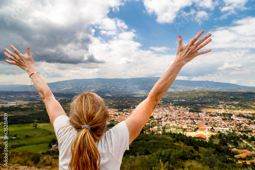 Woman hiker with arms raised looks at the Colombian town Villa de Leyva from the top of a hill