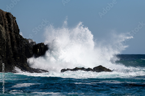 A large teal green color massive rip curl wave as it reaches the rocky shore. There's white frothy water with a textured water spray blowing off the tip of the wave with a dark blue ocean.