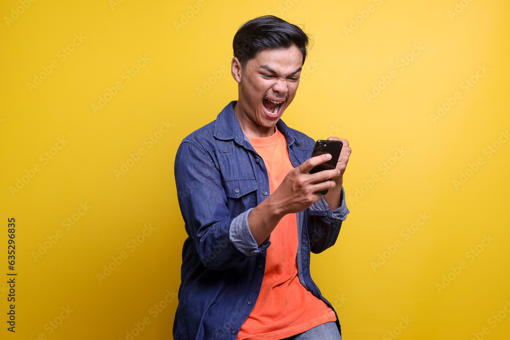 Asian man showing rage expression when looking to his cellular phone against yellow background an adult