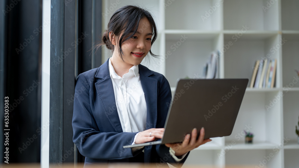 A beautiful Asian businesswoman using her laptop while standing by the window in her office. A beautiful Asian businesswoman using her laptop while standing by the window in her office.