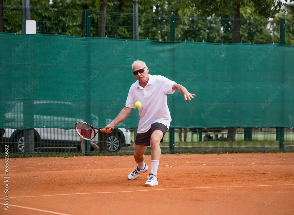 An elderly man playing tennis is hitting a forehand. The moving at camera. Open ground.