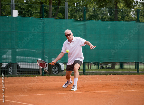 An elderly man playing tennis is hitting a forehand. The moving at camera. Open ground. © Дворецкая Таня