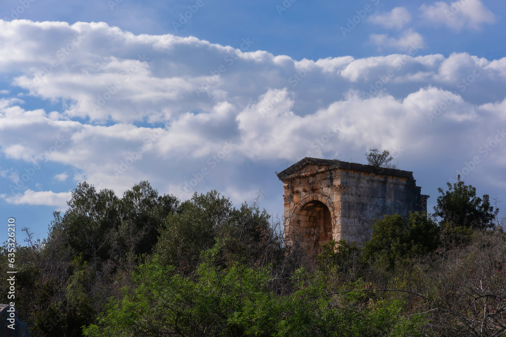 A Roman civilization artifact, located amidst the greenery in Anatolia, stands beautifully illuminated by the sun, casting its rays from the cloudy blue sky.