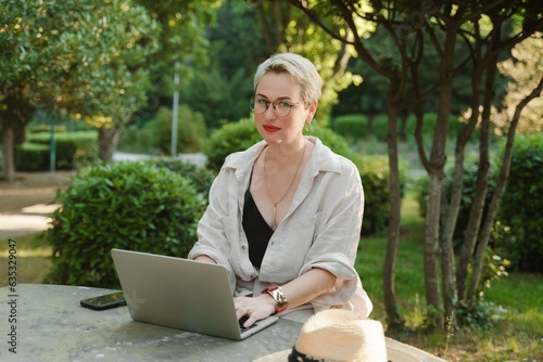 woman freelancer in glasses works at a computer at a white table in nature and spends her day productively