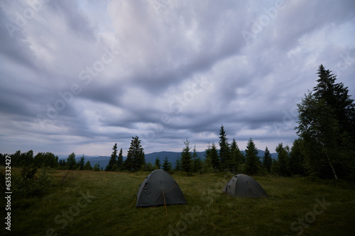 Panoramic view of mountain meadow with green grass  tourist tents and coniferous trees under cloudy sky. Dramatic sky with clouds over campsite in mountains. Camping and traveling concept.