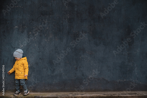 A little boy 2-3 years old in a yellow rubber raincoat against a gray loft wall. Lonely small figure on a dark background. Sad lost child. Mental health and autism concept