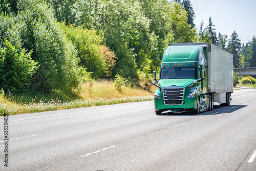 Green big rig bonnet long haul semi truck transporting cargo in dry van semi trailer driving on the wide highway road along the wild forest