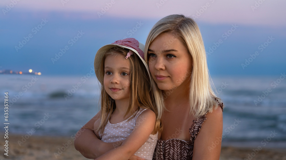 Mother and little daughter on beach of sea or ocean in evening at sunset. Summer vacation, travel, holiday. People outdoors